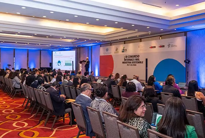 Audiencia atenta en el congreso internacional sobre industria sostenible, con un orador presentando en el escenario y pantallas mostrando gráficos y datos.