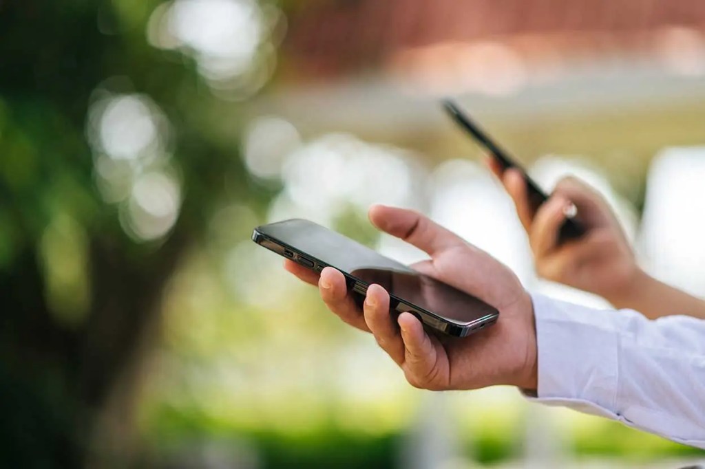 Una persona sostiene un teléfono móvil en una mano, mientras que otra persona sostiene un teléfono en la parte posterior, con un fondo natural desenfocado.
