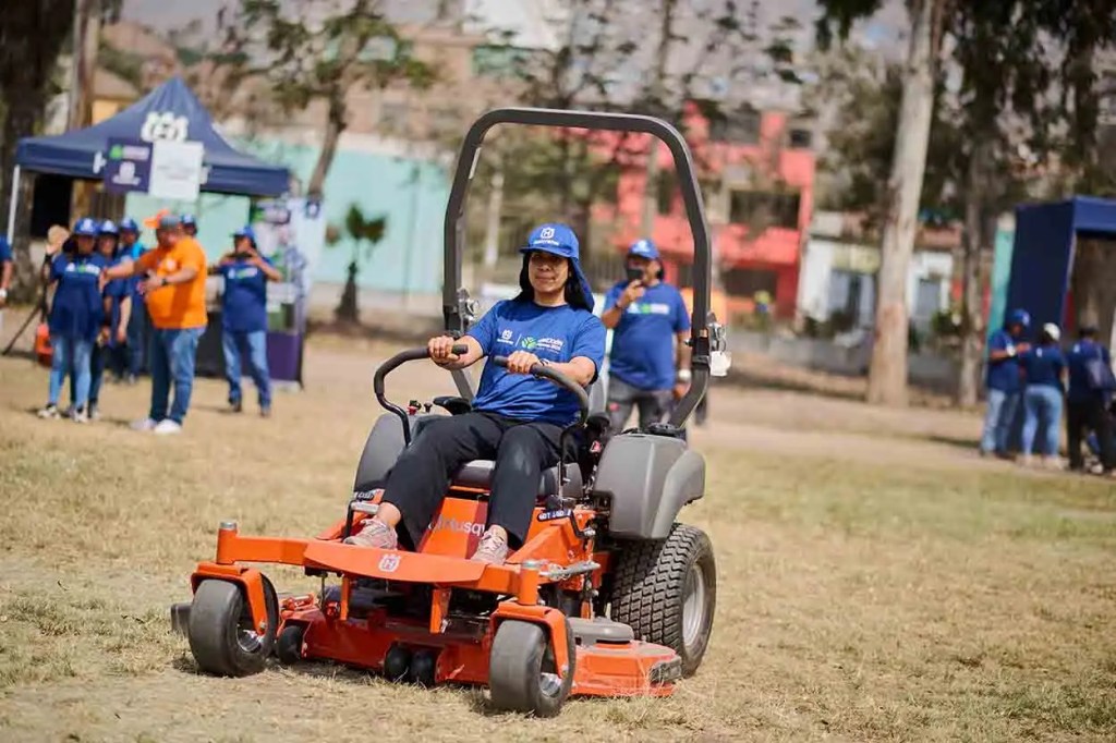 Una mujer maneja una cortadora de césped Husqvarna en un evento de jardinería al aire libre, con personas observando al fondo.