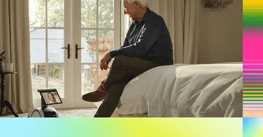 Un hombre mayor sentado en la cama interactuando con un robot que muestra una pantalla, en un entorno de hogar acogedor con luz natural entrando por la ventana.