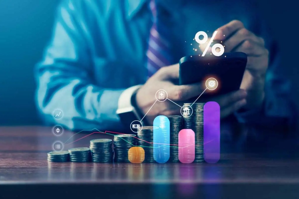 Un hombre en un traje sostiene un teléfono móvil mientras observa gráficos y monedas apiladas sobre una mesa, simbolizando el crecimiento financiero y la tecnología en finanzas.