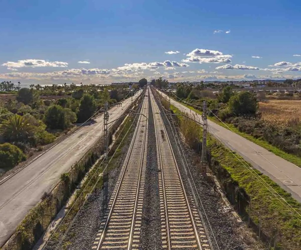 Vista de una vía de tren que se extiende hacia el horizonte, flanqueada por vegetación y una carretera a un costado, bajo un cielo parcialmente nublado.