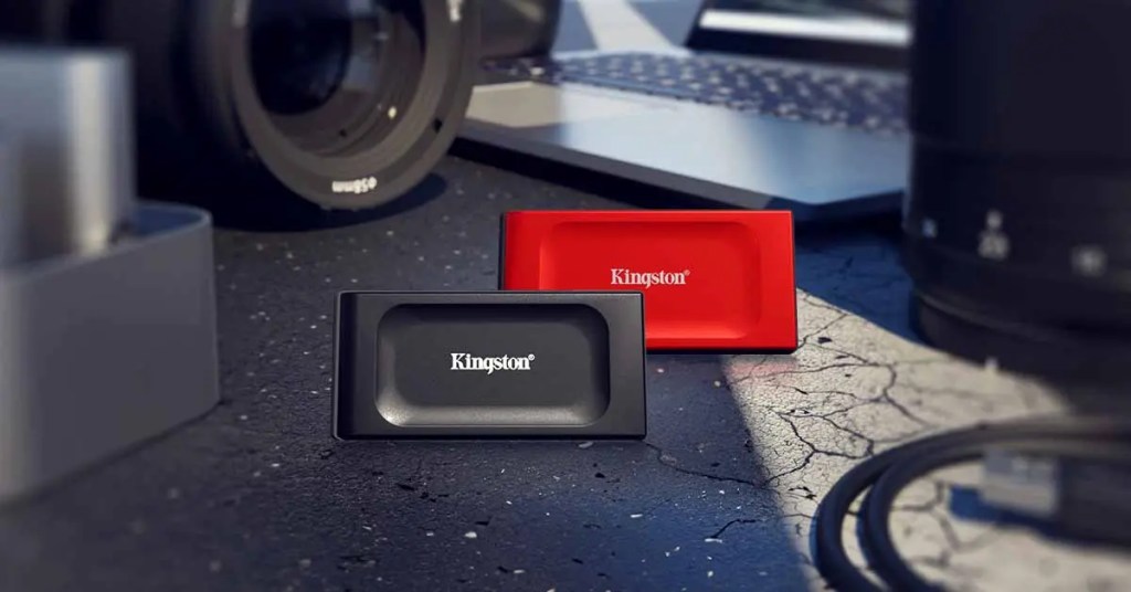 Dos unidades de almacenamiento Kingston, una negra y una roja, sobre una superficie de trabajo con cámara y otros dispositivos electrónicos de fondo.