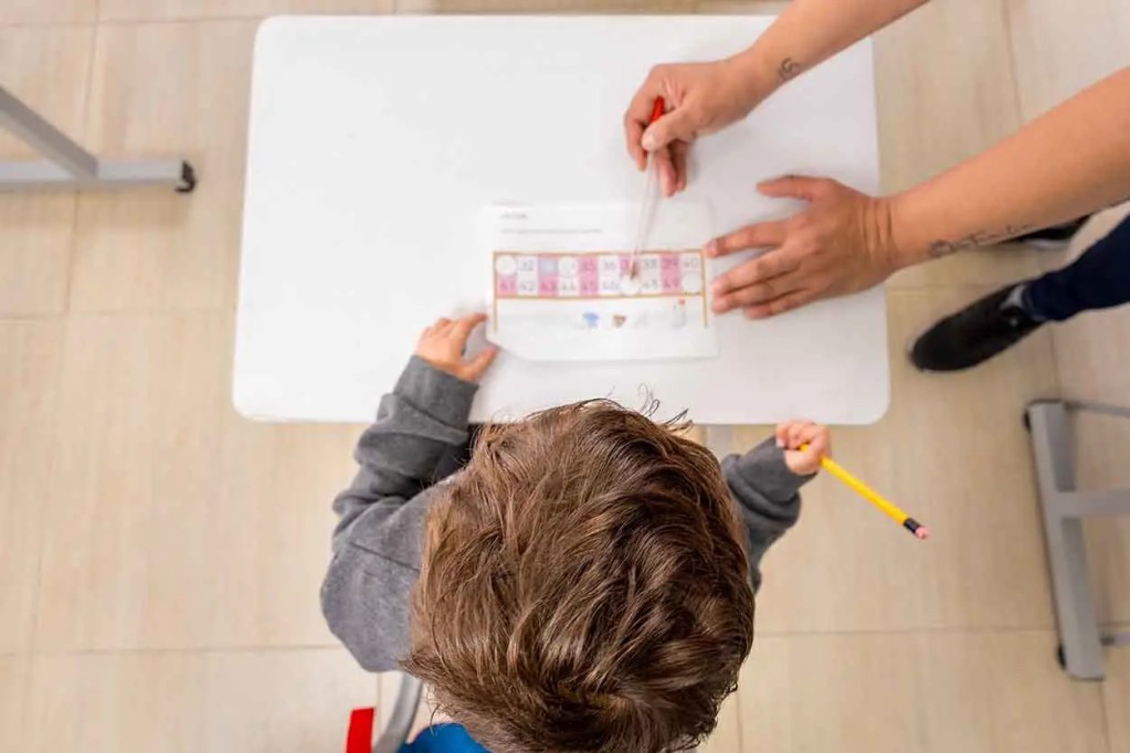 Un niño sentado en una mesa, observando una hoja de ejercicios de matemáticas mientras un adulto le señala con un lápiz rojo.