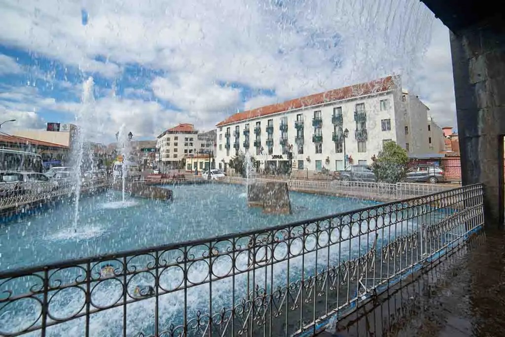 Vista de la fuente ornamental La Paccha en Cusco, con el edificio del hotel Xima Exclusive Cusco al fondo bajo un cielo nublado.