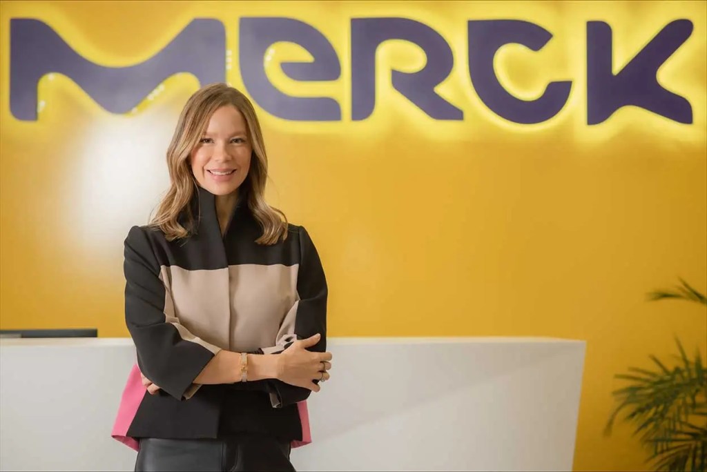 Mujer sonriente posando frente al logo de Merck en un ambiente de oficina.