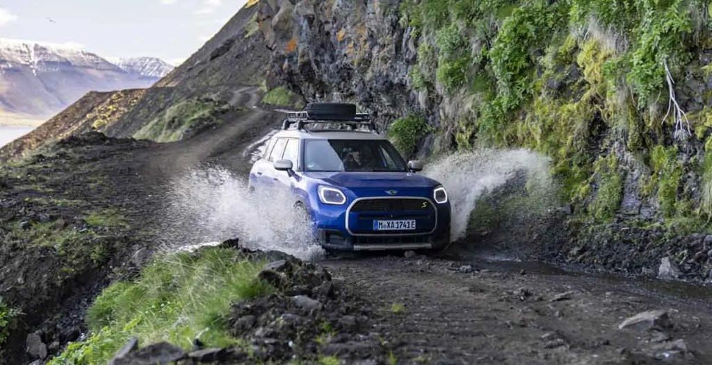 Un vehículo azul circulando por un camino de tierra, salpicando agua y rodeado de naturaleza montañosa.