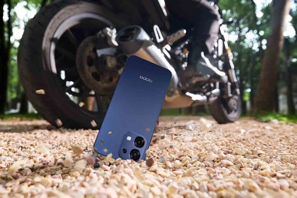 Un smartphone OPPO A6 Pro 5G de color azul se encuentra en el suelo, con una motocicleta al fondo. La imagen destaca el teléfono sobre un lecho de piedras en un entorno natural.