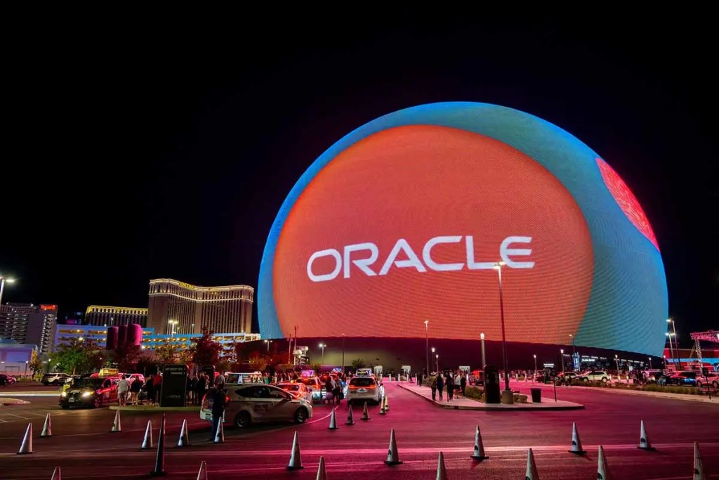 Exterior del evento Oracle AI World 2025 en Las Vegas, con un gran display iluminado que muestra el logo de Oracle, vehículos estacionados y personas en el área.