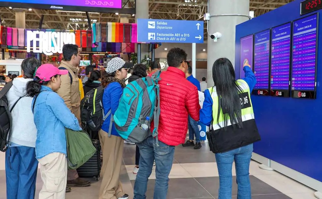Pasajeros en el Aeropuerto Internacional Jorge Chávez observando la pantalla de salidas mientras una trabajadora les indica información.