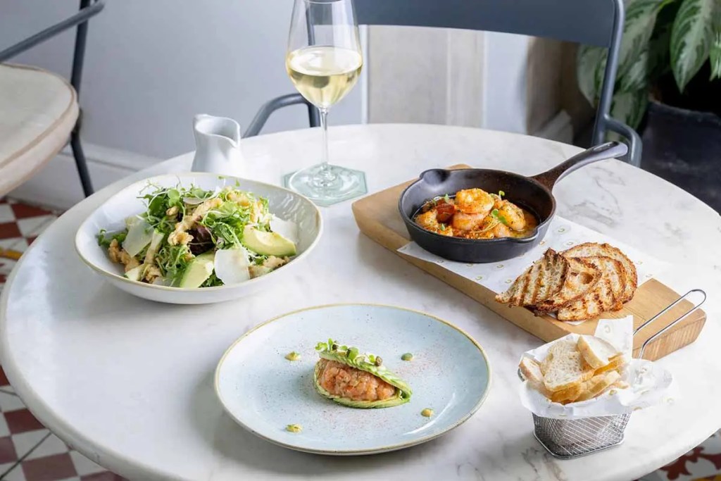 Una mesa con una ensalada verde, un plato de mariscos en una sartén de hierro fundido, tostadas y pan en una canasta, y una copa de vino blanco.