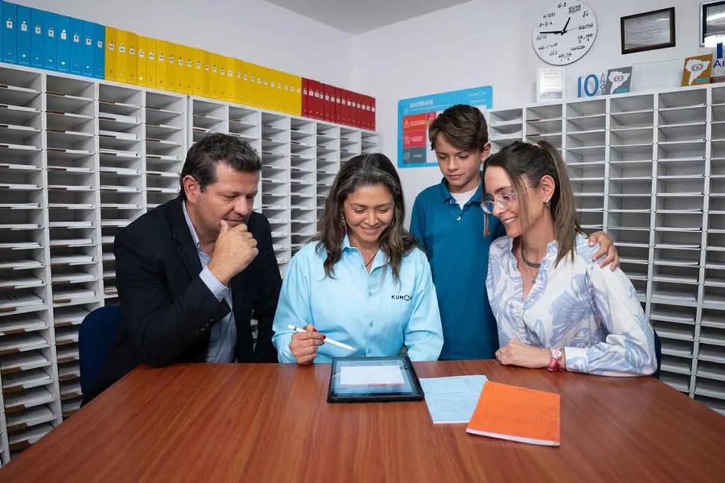 Un grupo de cuatro personas, incluyendo un niño, se reúne alrededor de una tablet en una mesa. Hay documentos y cuadernos en la mesa, y un fondo de estanterías con archivos de colores.