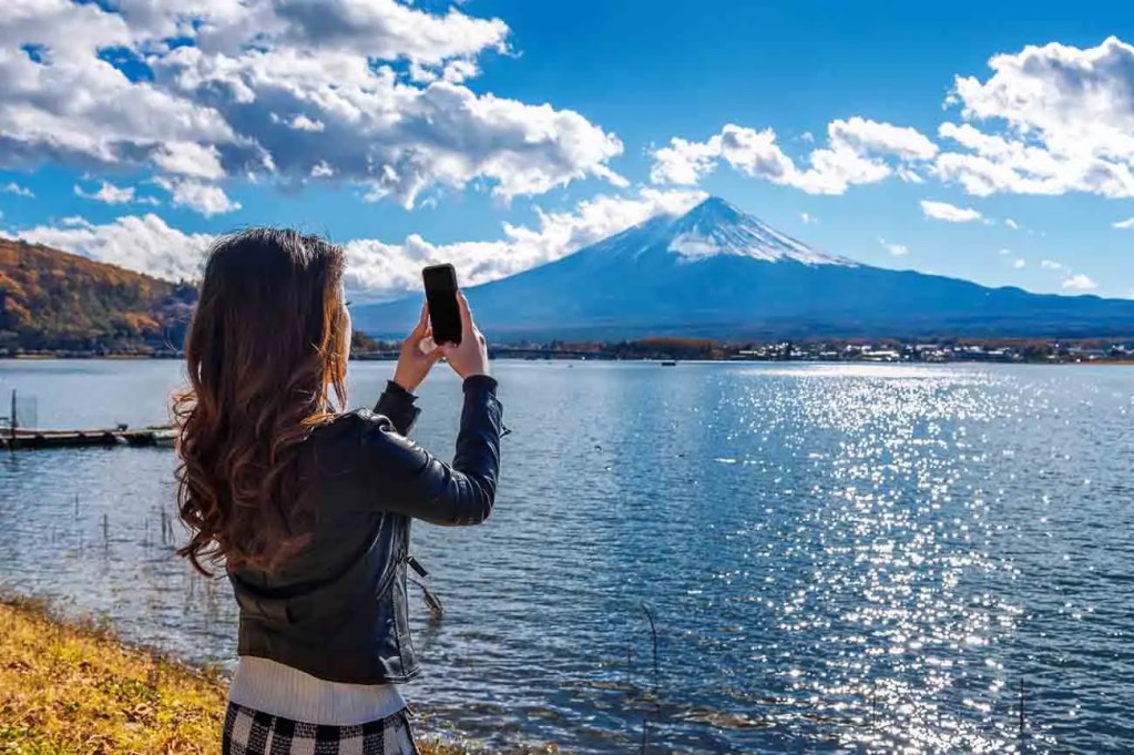 Una mujer de pie junto a un lago, tomando una foto del paisaje con su teléfono móvil. Al fondo se observa el monte Fuji, rodeado de nubes y un cielo despejado.