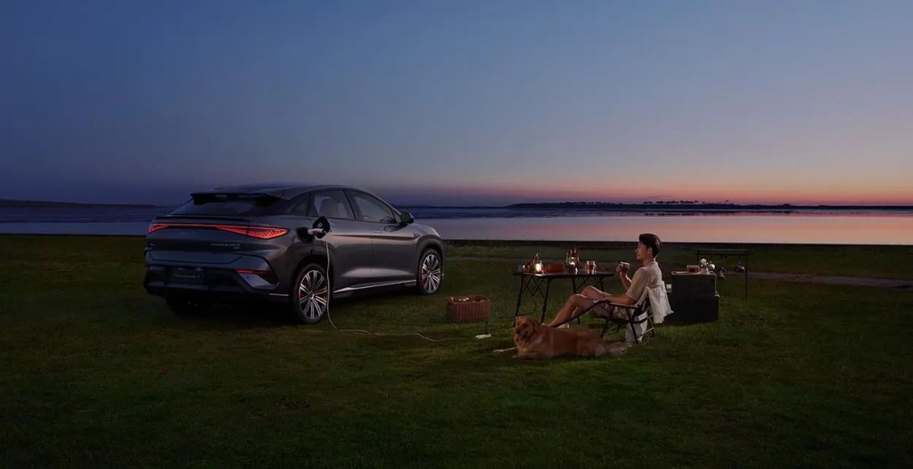 Un vehículo eléctrico de BYD estacionado en un paisaje al atardecer, mientras una persona disfruta de un momento de relajación al aire libre con un perro a su lado.