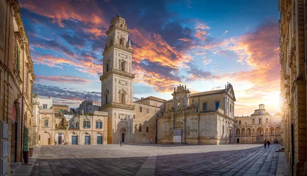 Vista panorámica de la Piazza Sant'Oronzo en Lecce, Italia, con un cielo colorido al atardecer y edificios de arquitectura barroca.