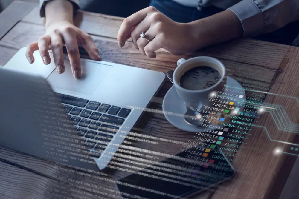 Una persona utilizando una computadora portátil en una mesa de madera, con una taza de café al lado. La pantalla muestra datos digitales y un teclado con manos en uso.