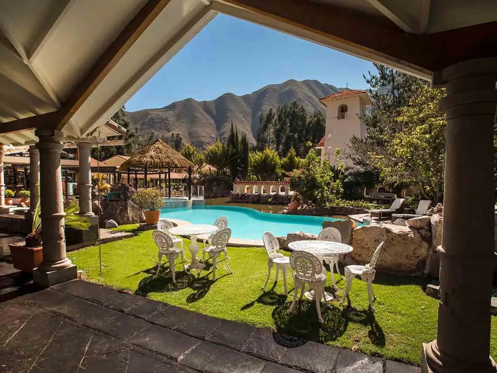 Vista de un área al aire libre en un hotel con piscina, rodeada de montañas y vegetación, equipada con mesas y sillas blancas.
