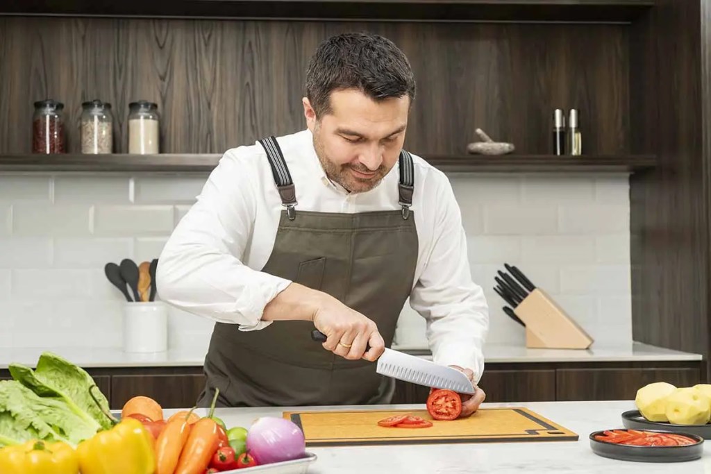 Un chef en un entorno de cocina, cortando un tomate sobre una tabla, rodeado de una variedad de vegetales frescos y utensilios de cocina.