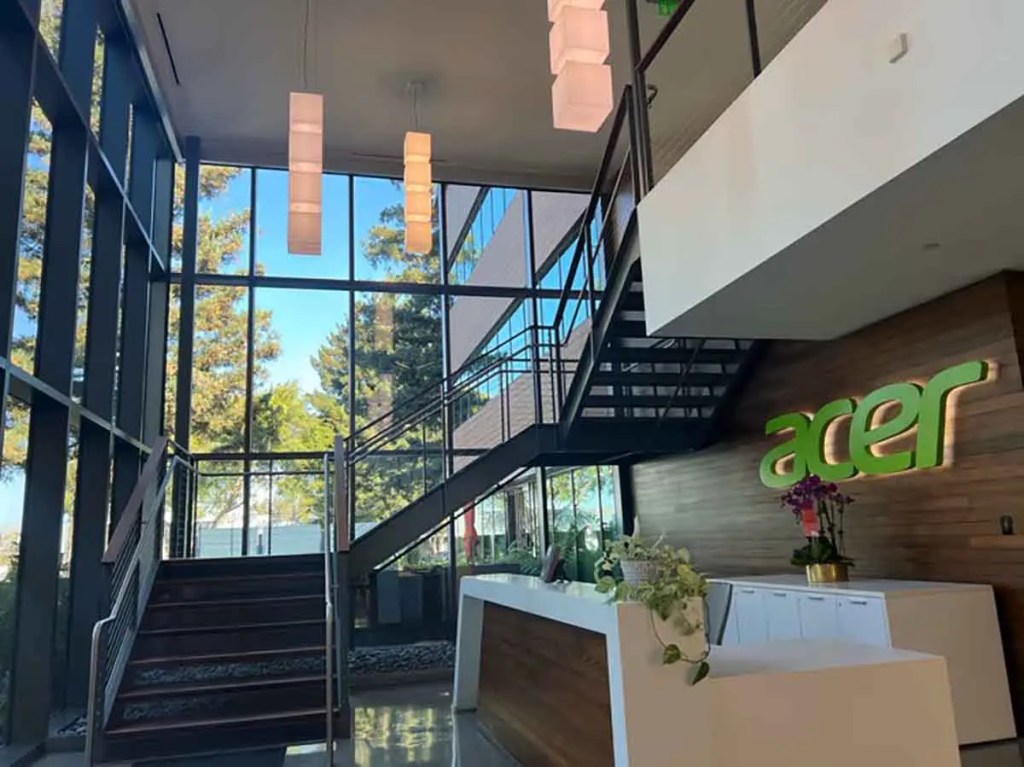 Interior de la sede de Acer con un diseño moderno, escaleras y un mostrador de recepción decorado con plantas, iluminado por ventanas grandes que muestran árboles al exterior.