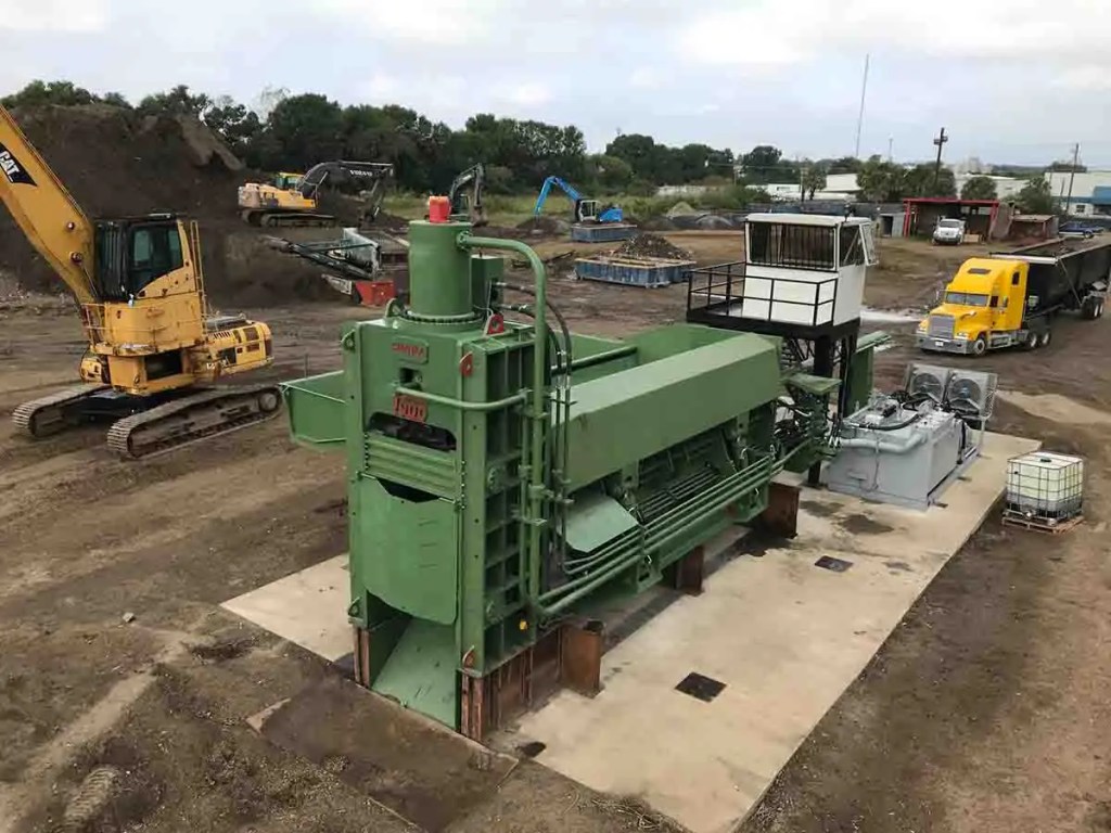 Vista de maquinaria industrial en un área de construcción, incluyendo una fragmentadora verde, una cabina de control, y equipos móviles en el fondo.