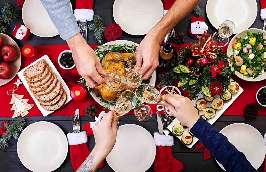 Mesa festiva decorada para una celebración navideña, con platos de comida, brindis con copas y elementos decorativos como manzanas, velas y adornos navideños.