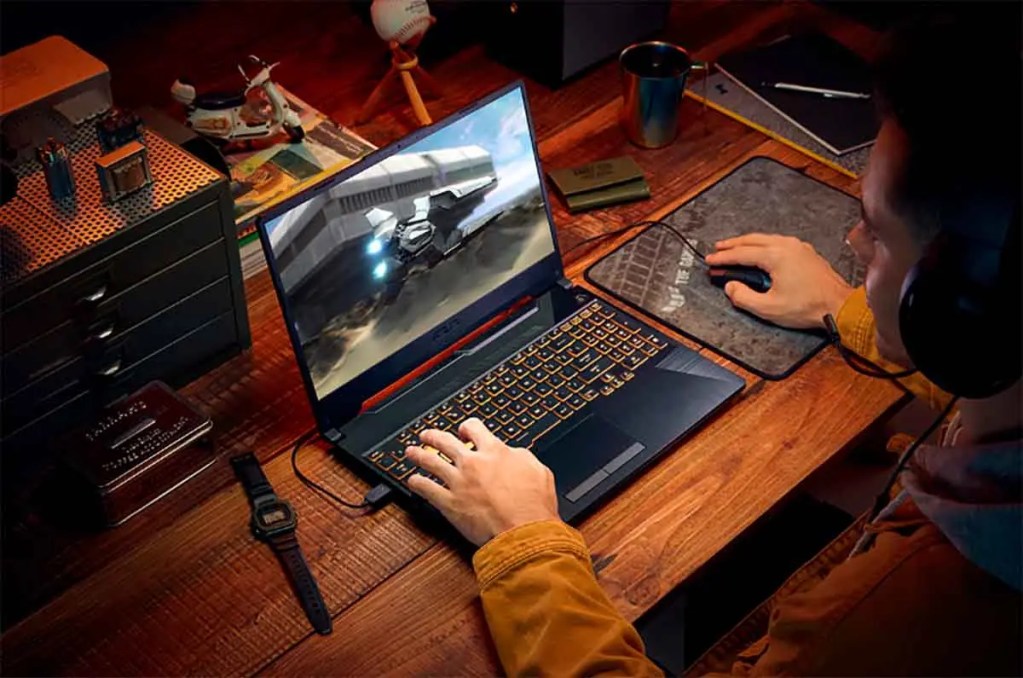 Persona jugando en una laptop gamer en un escritorio de madera, con un juego de alta acción visible en la pantalla. Se observan accesorios de escritorio como un reloj y un mousepad.