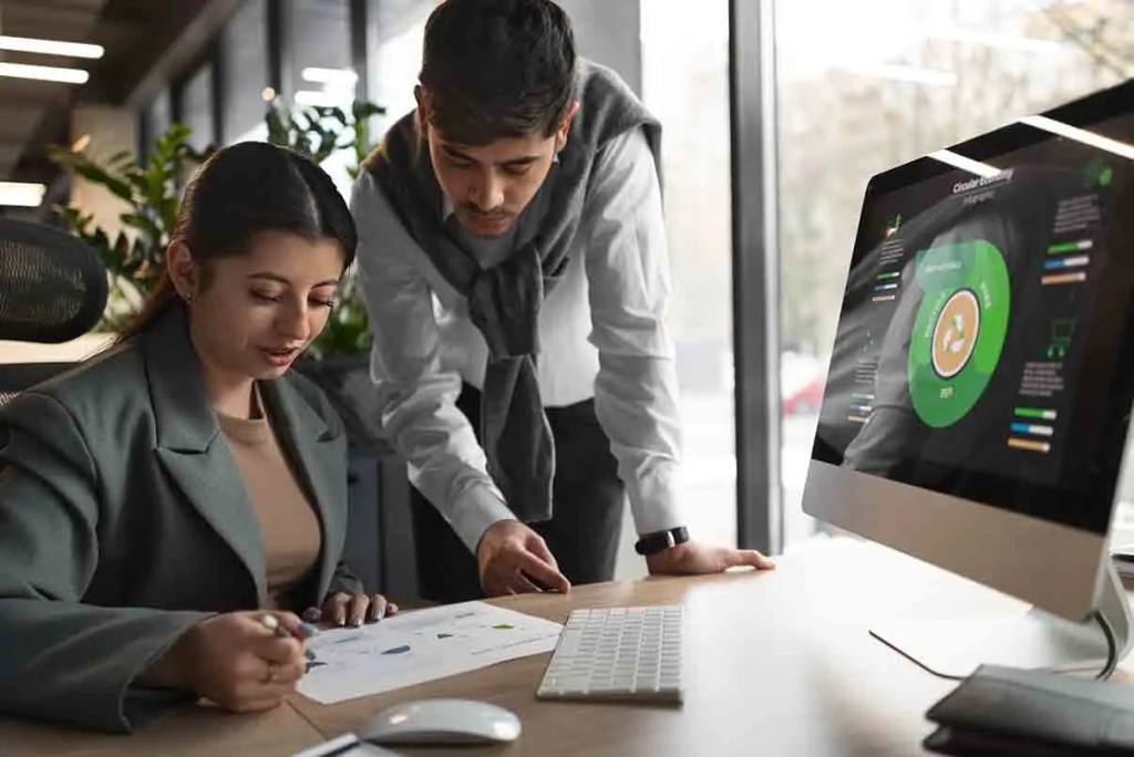 Una mujer y un hombre revisando gráficos en una computadora mientras la mujer toma notas en una hoja de papel en una oficina moderna.
