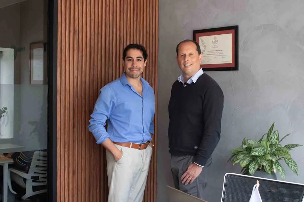 Dos hombres de pie frente a una pared de madera y una planta, en un entorno de oficina, discutendo sobre la financiera tecnológica Capitalia.