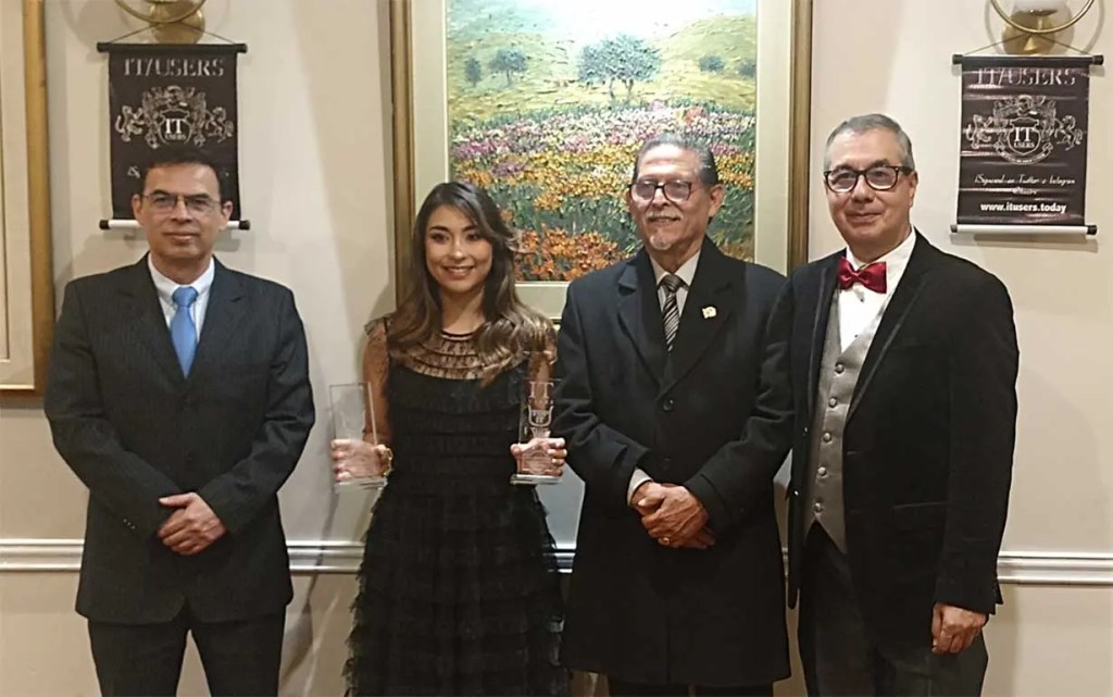 Cuatro personas sonríen en una ceremonia de premiación, sosteniendo trofeos. Al fondo, una pintura de un paisaje floral.