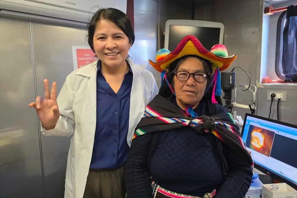 Una trabajadora de salud sonriente junto a una mujer indígena que lleva un sombrero colorido en un ambiente de atención médica.