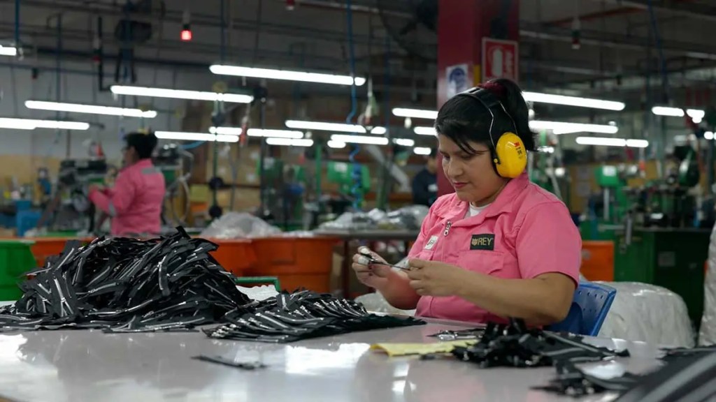 Trabajadora en planta de producción de Corporación Rey, organizando piezas en un ambiente industrial. Se observan maquinaria y otros empleados en el fondo.