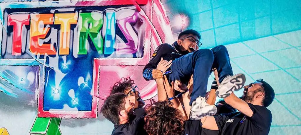 Un grupo de personas celebra levantando a un jugador en un evento de Red Bull Tetris®, con un fondo colorido que muestra el logo del juego.