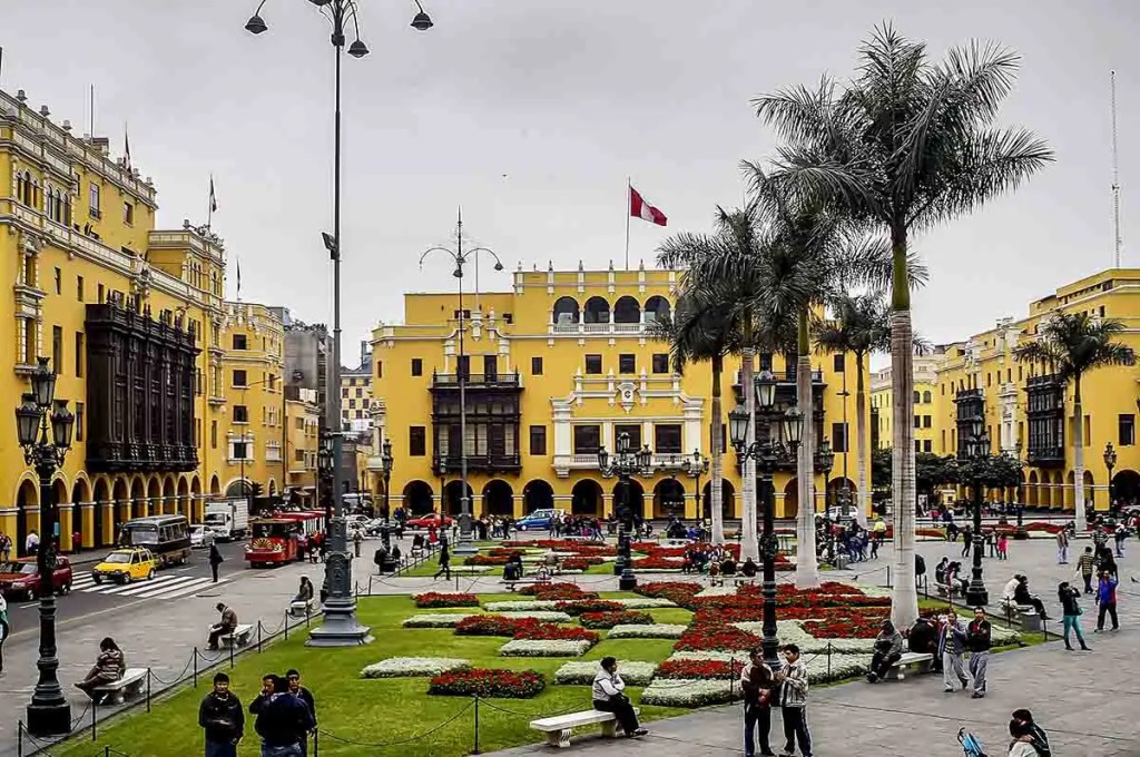 Vista de la Plaza Mayor de Lima, Perú, con edificios históricos de color amarillo, palmeras y un jardín bien cuidado, rodeado de visitantes y vehículos.