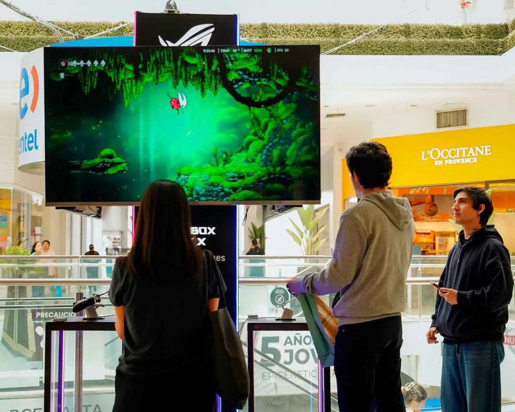 Tres personas interactuando en un espacio de gaming en un centro comercial, observando un juego proyectado en una pantalla grande con imágenes de un entorno verde y un personaje volador.