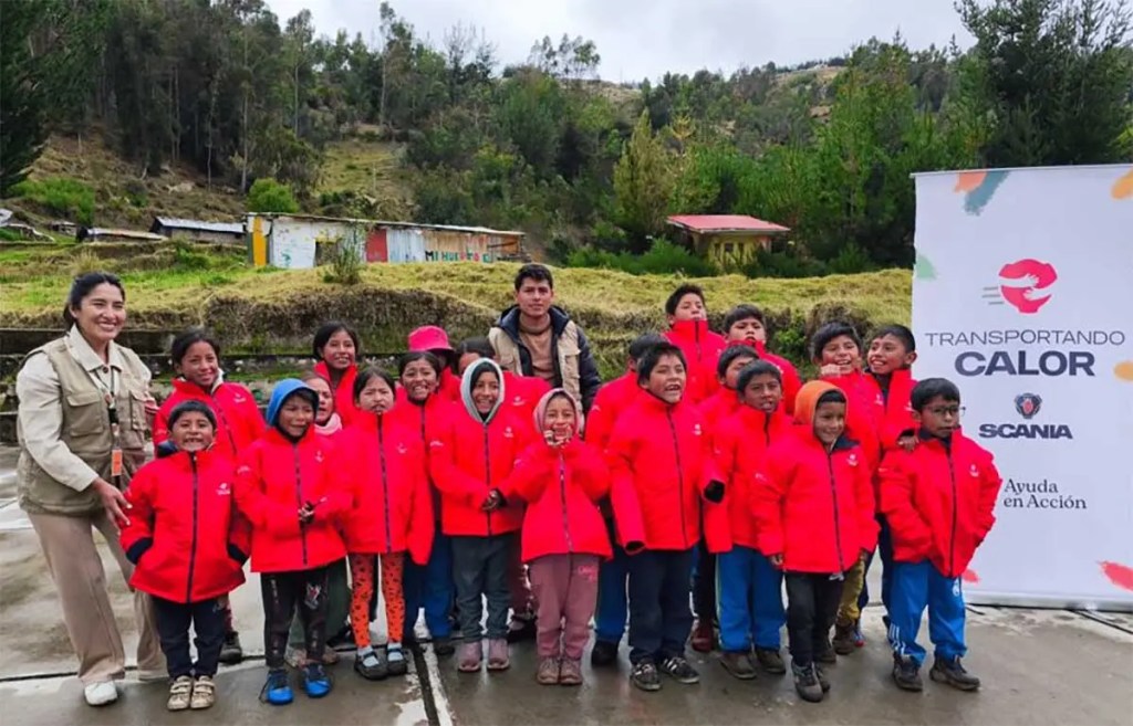 Grupo de niños y niñas de comunidades altoandinas en Apurímac y Cusco, vistiendo casacas térmicas rojas, junto a colaboradores de Scania Perú y un cartel del programa 'Transportando Calor'.