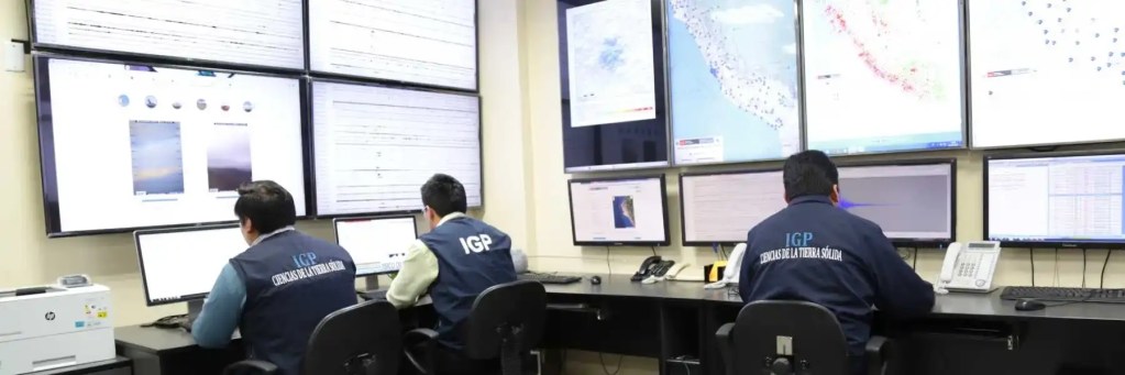 Tres operadores trabajando en una sala de monitoreo sísmico del Instituto Geofísico del Perú, con múltiples pantallas mostrando datos sísmicos y mapas.
