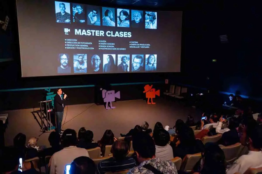 Evento de cine con un orador en el escenario presentando las clases magistrales junto a una proyección en pantalla sobre producción cinematográfica, con una audiencia visible en el fondo.