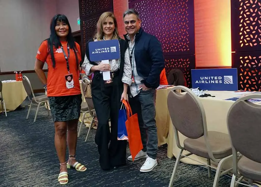 Tres personas posando en un evento, sosteniendo un cartel de United Airlines. Fondo con decoración del evento y mesas.