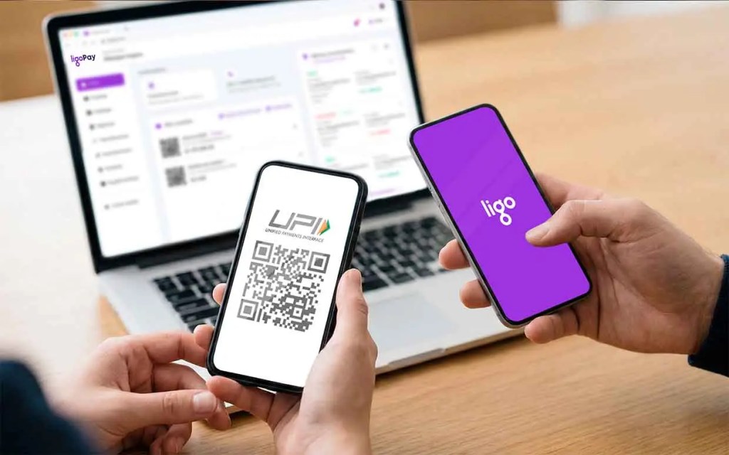 Una persona sostiene dos teléfonos móviles, uno mostrando un código QR para UPI (Interfaz Unificada de Pagos) y el otro con el logo de Ligo, sobre una mesa que también tiene una computadora portátil con una interfaz de software visible.