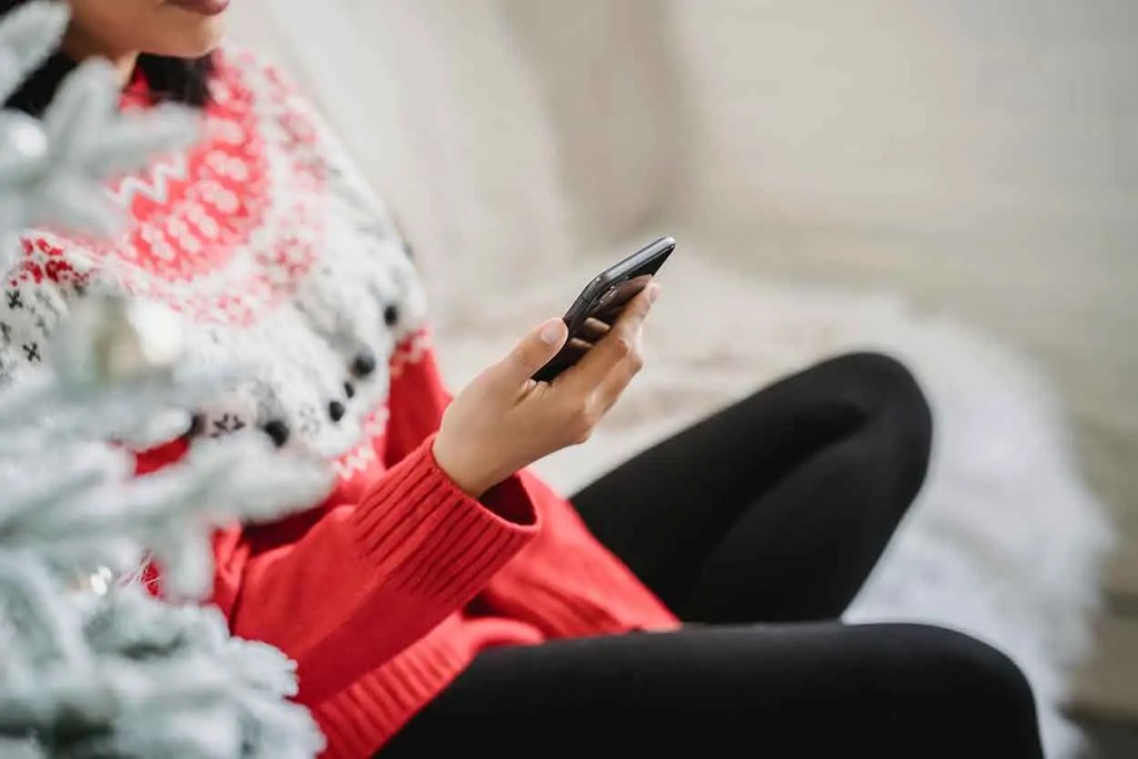 Mujer sentada con un suéter rojo y blanco, usando un teléfono móvil, en un entorno navideño con un árbol decorado al fondo.