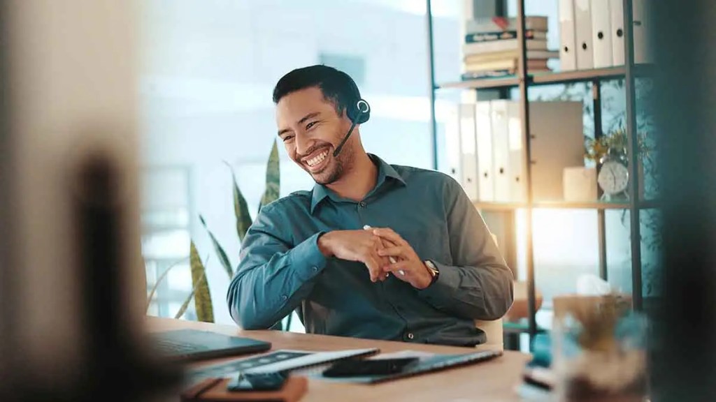 Hombre sonriendo mientras trabaja con auriculares en una oficina moderna, rodeado de plantas y estanterías.