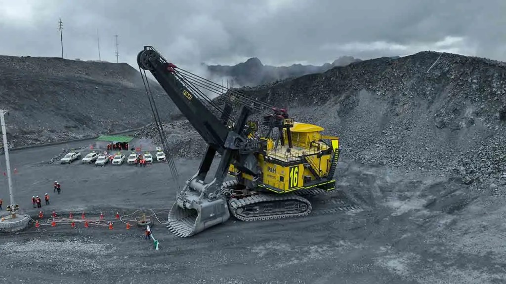Excavadora amarilla en una cantera, con trabajadores y vehículos en el fondo bajo un cielo nublado.
