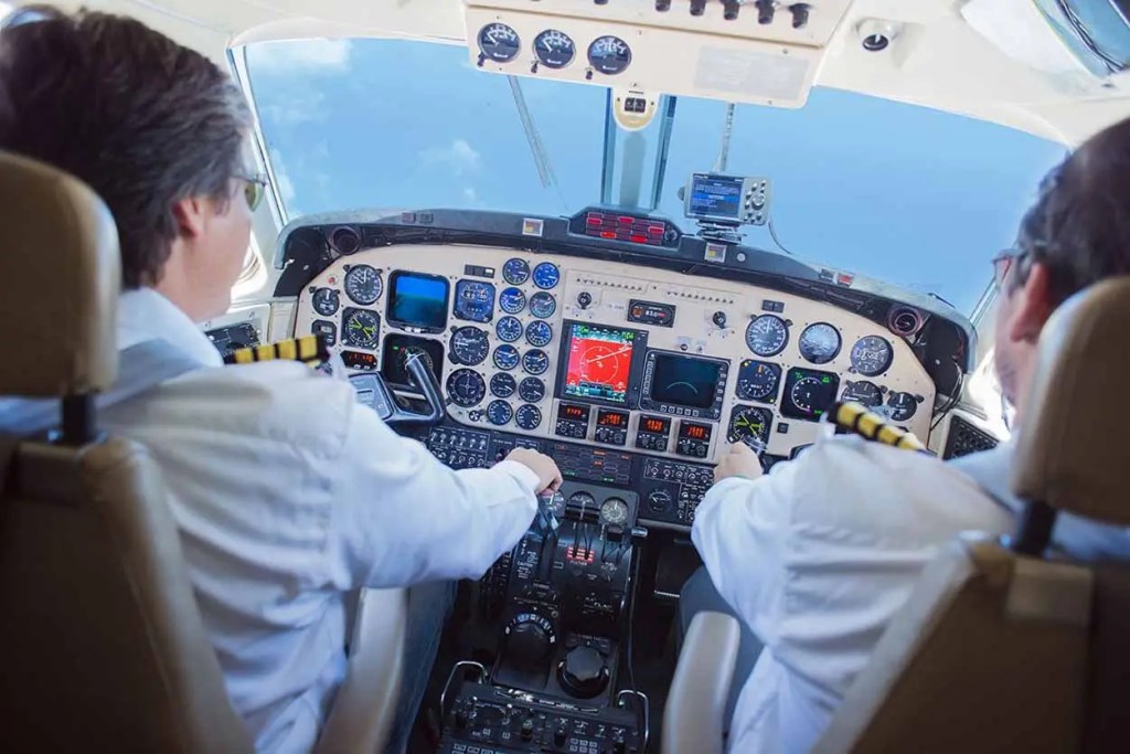 Cabina de avión con dos pilotos frente a los controles y pantallas de instrumentos.
