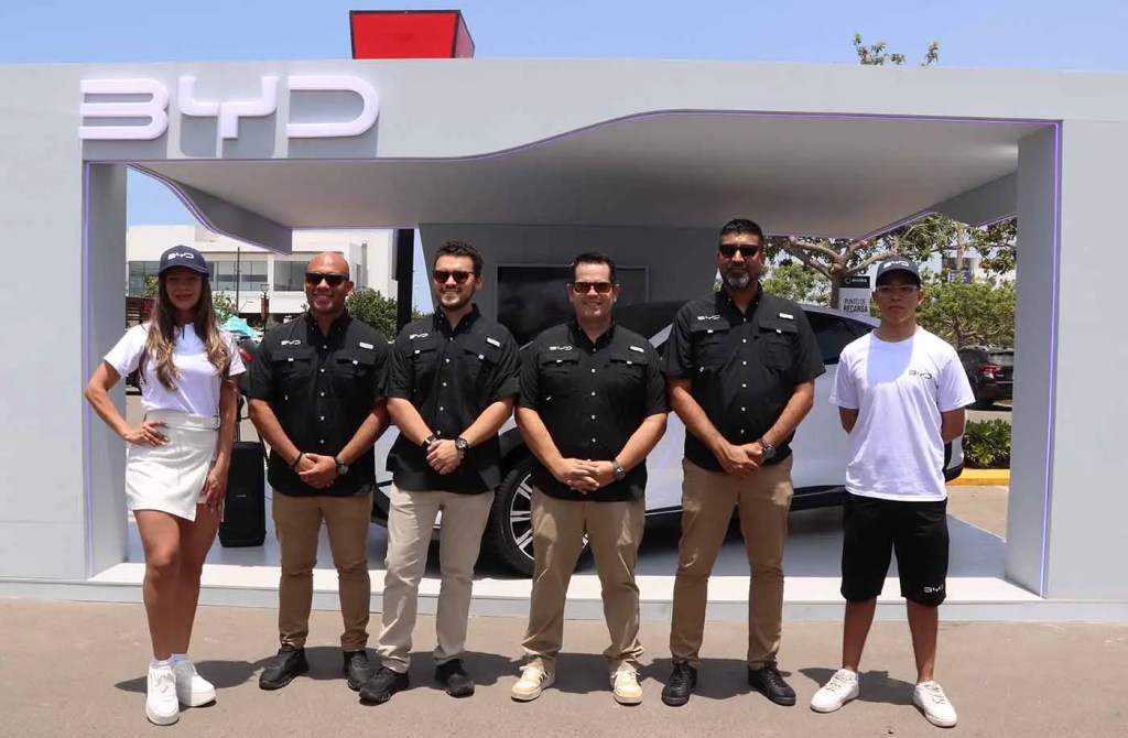 Grupo de seis personas posando frente a un stand de BYD, con vehículos eléctricos de fondo. La mujer a la izquierda lleva una camiseta blanca y una falda, mientras que los hombres usan camisas negras. El ambiente es soleado y al aire libre.