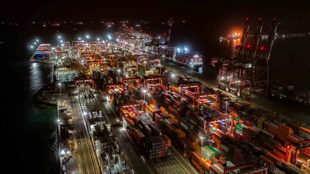 Vista aérea de un puerto con contenedores iluminados durante la noche, mostrando grúas y una gran cantidad de mercancía.