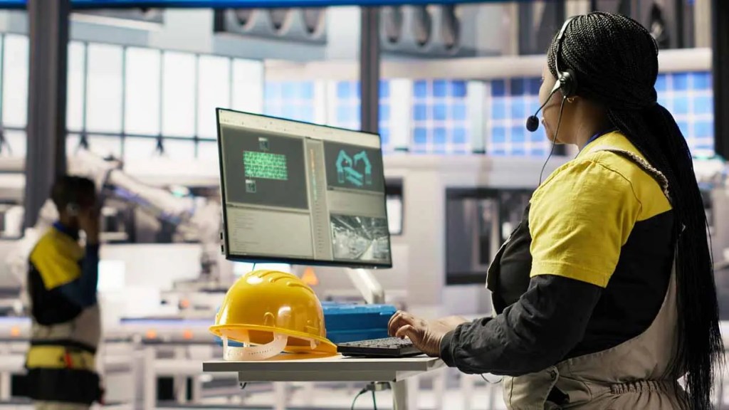 Mujer usando auriculares y operando una computadora en un entorno industrial con un casco de seguridad en la mesa.