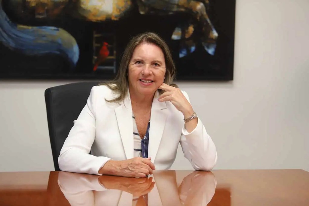 Mujer sonriendo sentada en una mesa de oficina, con cabello largo y chaqueta blanca, frente a un fondo oscuro con arte.