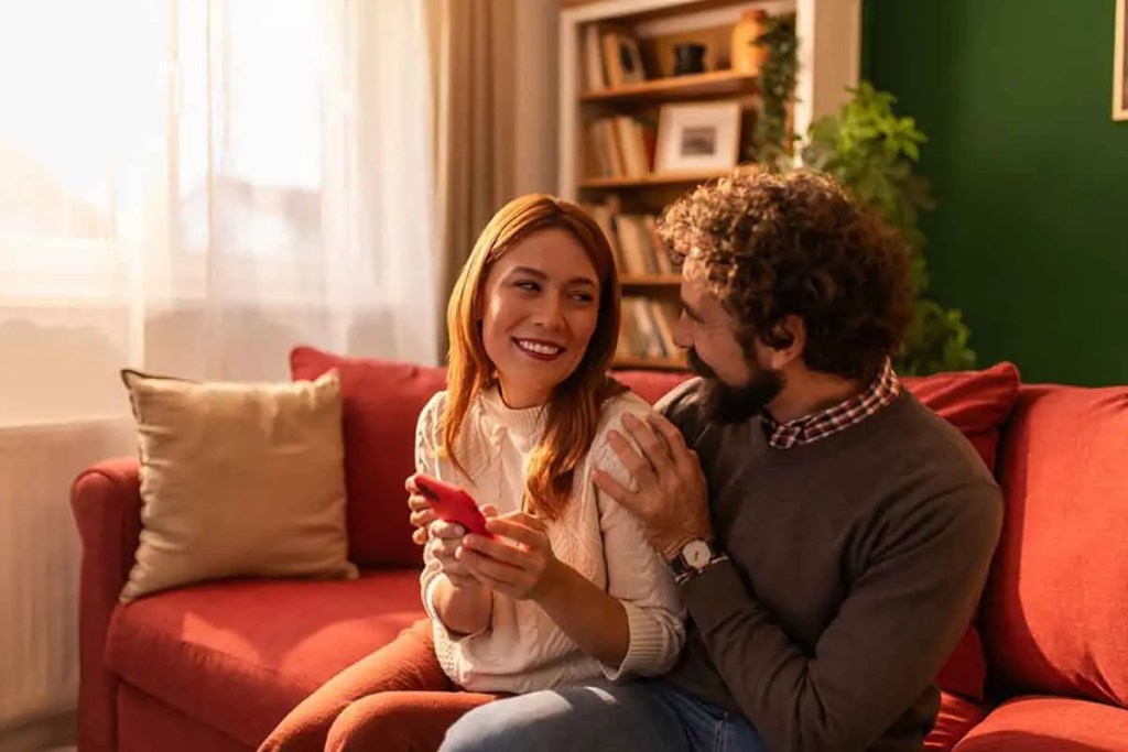 Una pareja sonriente sentada en un sofá, compartiendo un momento íntimo mientras miran un teléfono móvil.