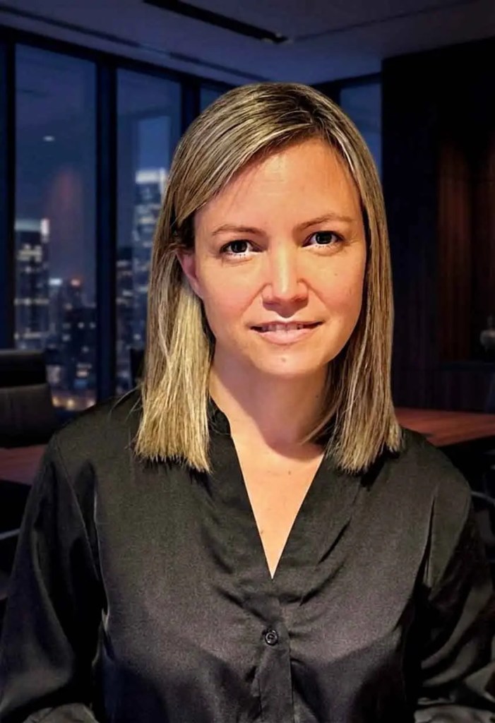 Mujer de cabello rubio y liso vestida con una camisa negra, posando en un entorno de oficina moderna con vistas a la ciudad de noche.