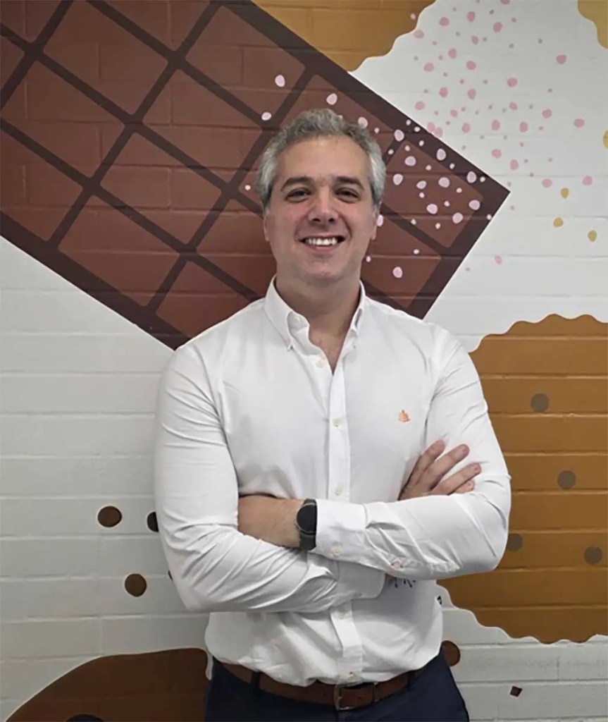 Hombre sonriendo con camisa blanca de pie frente a una pared decorada con chocolate y galletas.